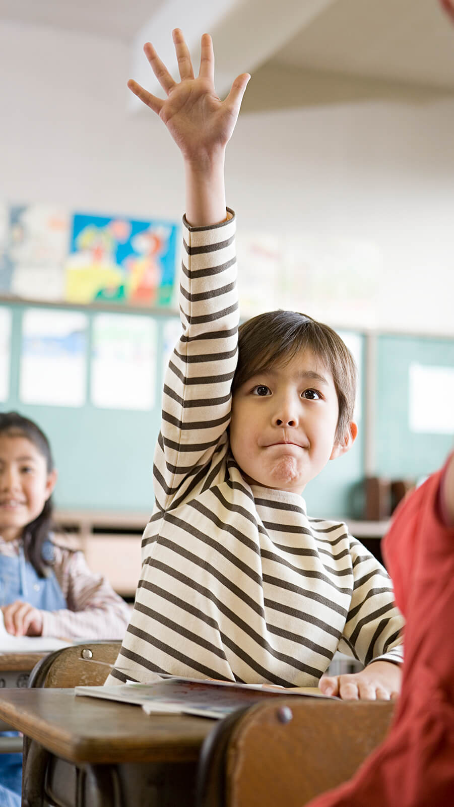 Young asian boy sitting in his classroom desk eagerly raising his hand.