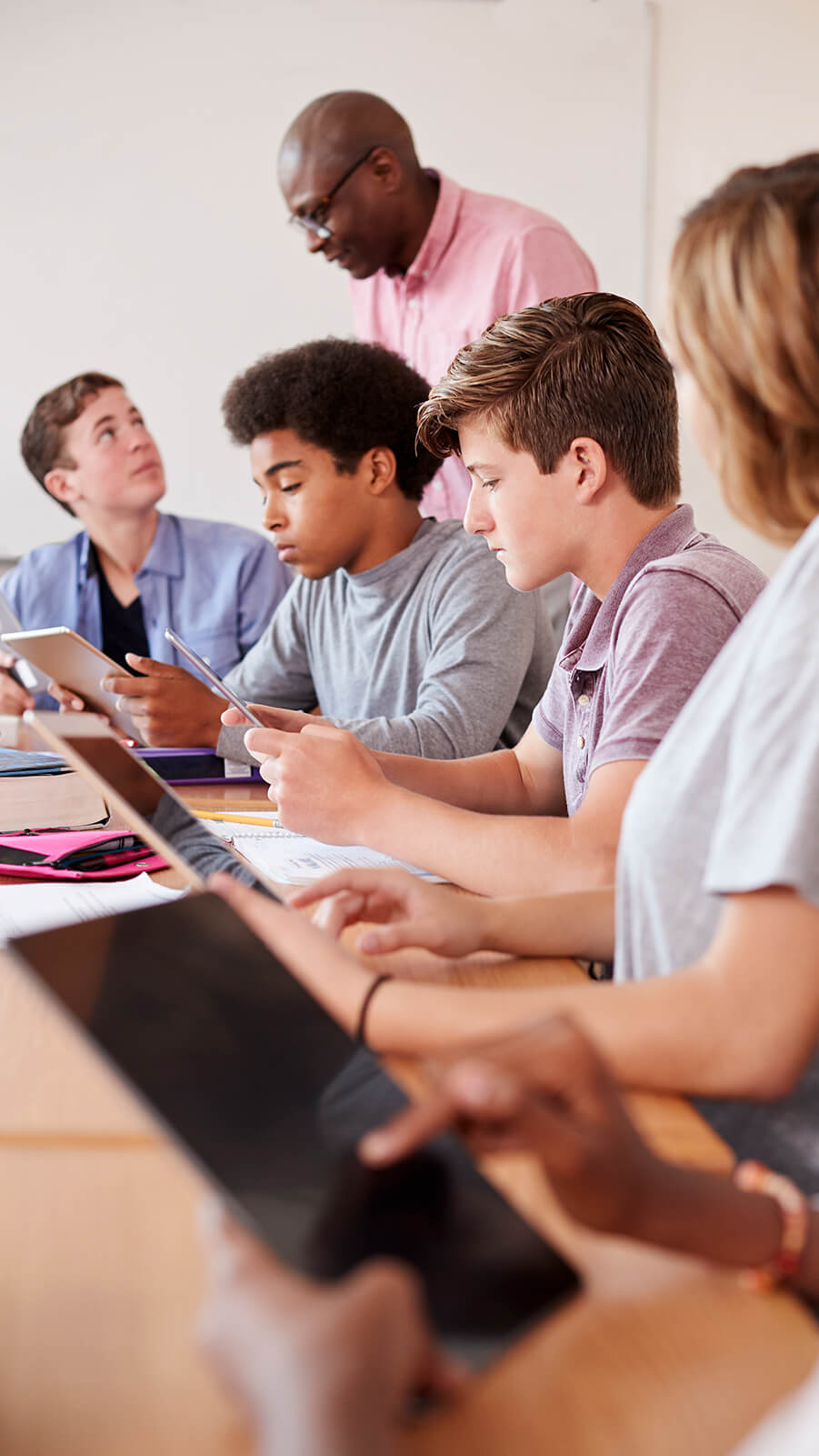 Four teenage students using tablets and a male instructor talking to one of them.