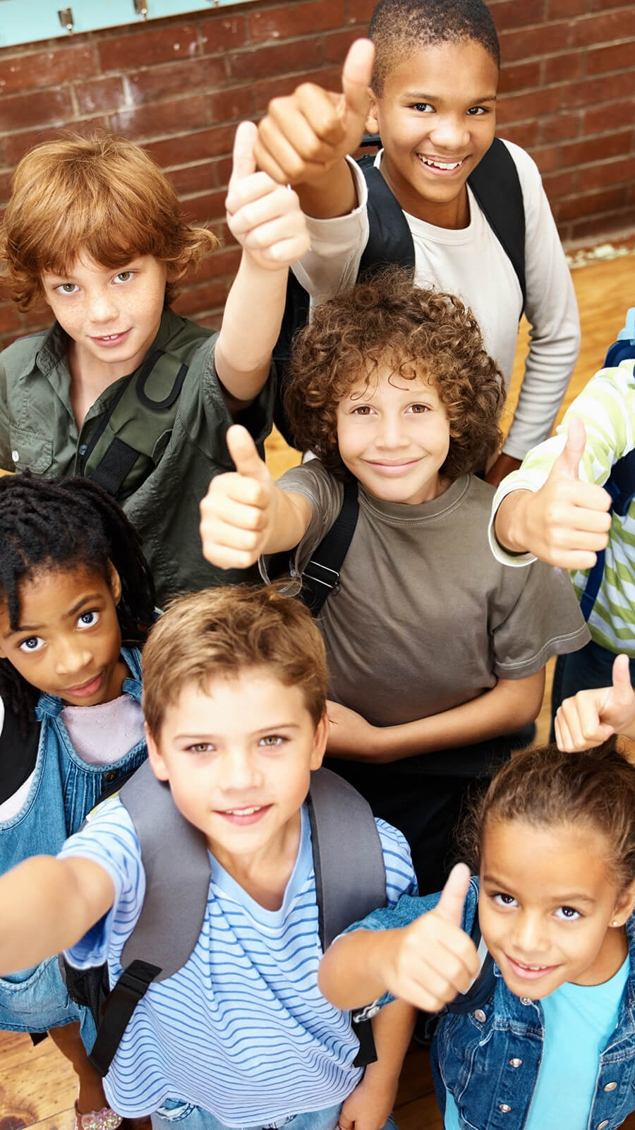 Group of male and female elementary school students wearing backpacks and holding thumbs up signs to the camera.